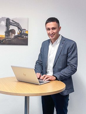 Ein Leyrer + Graf Mitarbeiter steht an einem Tisch und arbeitet am Laptop und lächelt dabei in die Kamera