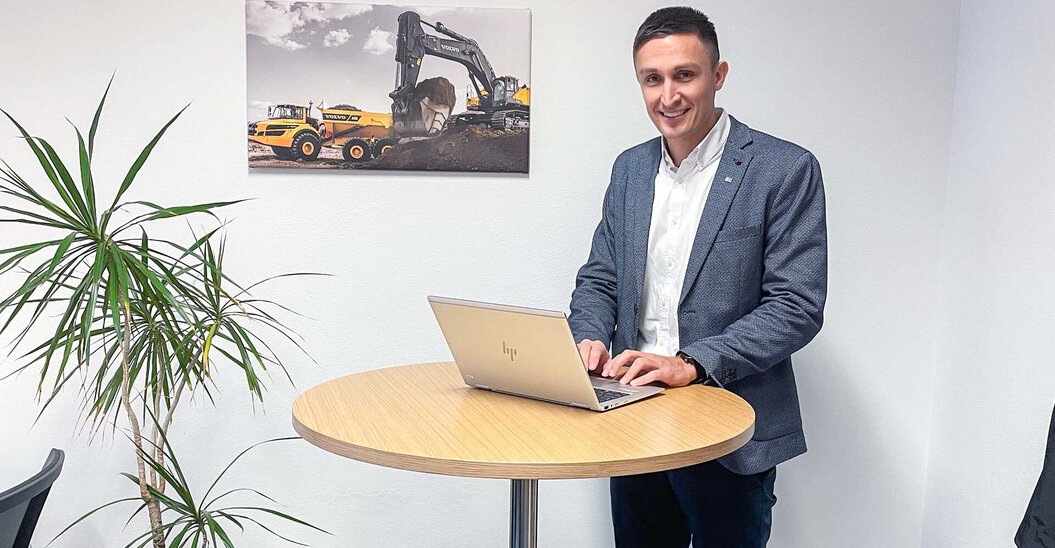 Ein Leyrer + Graf Mitarbeiter steht an einem Tisch und arbeitet am Laptop und lächelt dabei in die Kamera