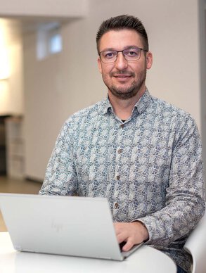 Leyrer + Graf Mitarbeiter sitz mit seinem Laptop an einem kleinen Tisch und lächelt dabei in die Kamera