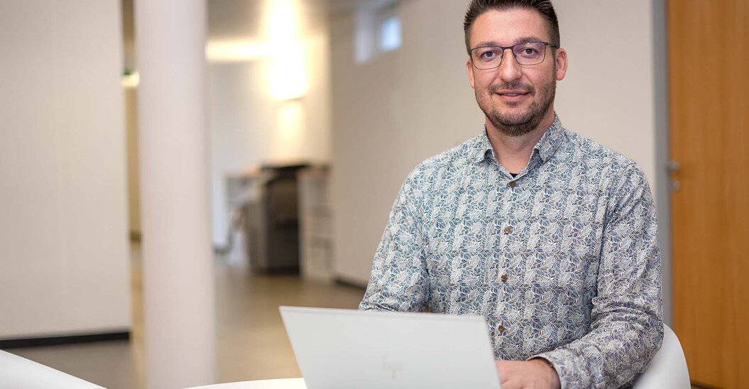 Leyrer + Graf Mitarbeiter sitz mit seinem Laptop an einem kleinen Tisch und lächelt dabei in die Kamera