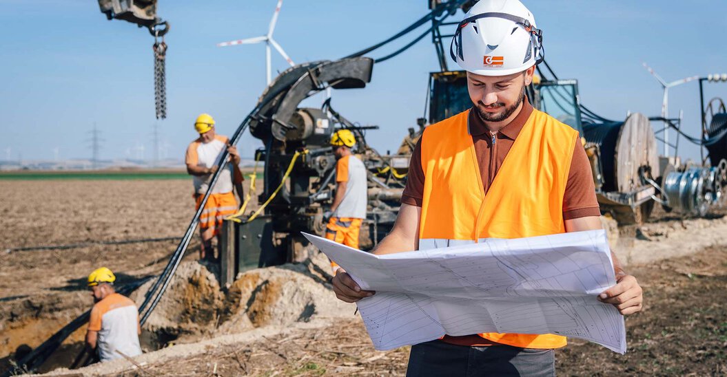 Ein Leyrer + Graf Mitarbeiter steht auf einer Baustelle und blickt auf einen Bauplan im Hintergrund ist ein Kabelpflug zu sehen