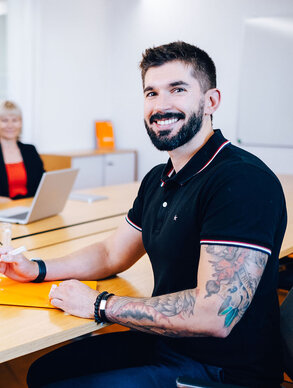 Leyrer + Graf Mitarbeiter blickt in die Kamera und lächelt im Hintergrund sind mehrere Kollegen in einer Besprechung zu sehen