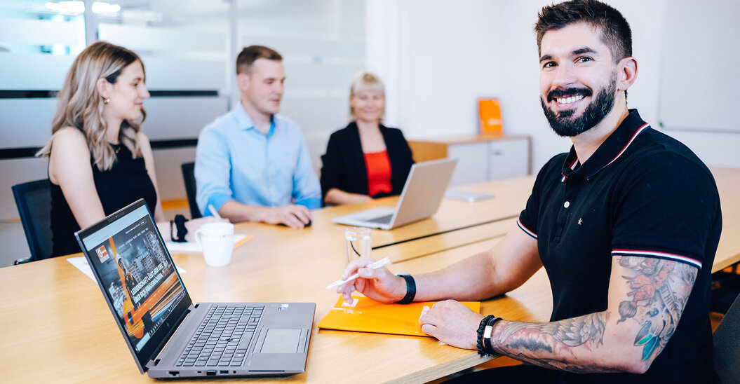 Leyrer + Graf Mitarbeiter blickt in die Kamera und lächelt im Hintergrund sind mehrere Kollegen in einer Besprechung zu sehen
