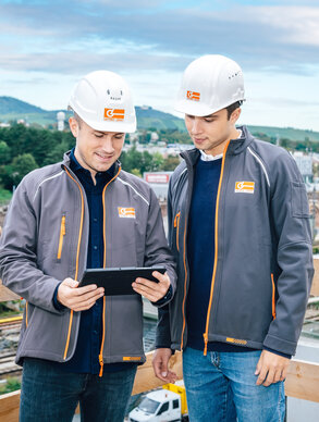 Zwei Leyrer + Graf Mitarbeiter besprechen einen Plan am Tablet im Hintergrund ist eine Baustelle und ein grüner Berg zu sehen