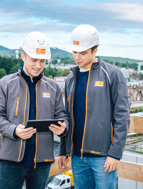 Zwei Leyrer + Graf Mitarbeiter besprechen einen Plan am Tablet im Hintergrund ist eine Baustelle und ein grüner Berg zu sehen