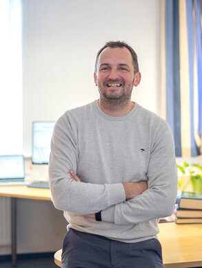 Leyrer + Graf Mitarbeiter lehnt an seinem Tisch mit verschränkten Händen und lächelt in die Kamera