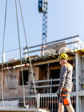 Ein Leyrer + Graf Mitarbeiter auf einer Baustelle neben ihm wird ein Betonelement mittels Kran hochgehoben